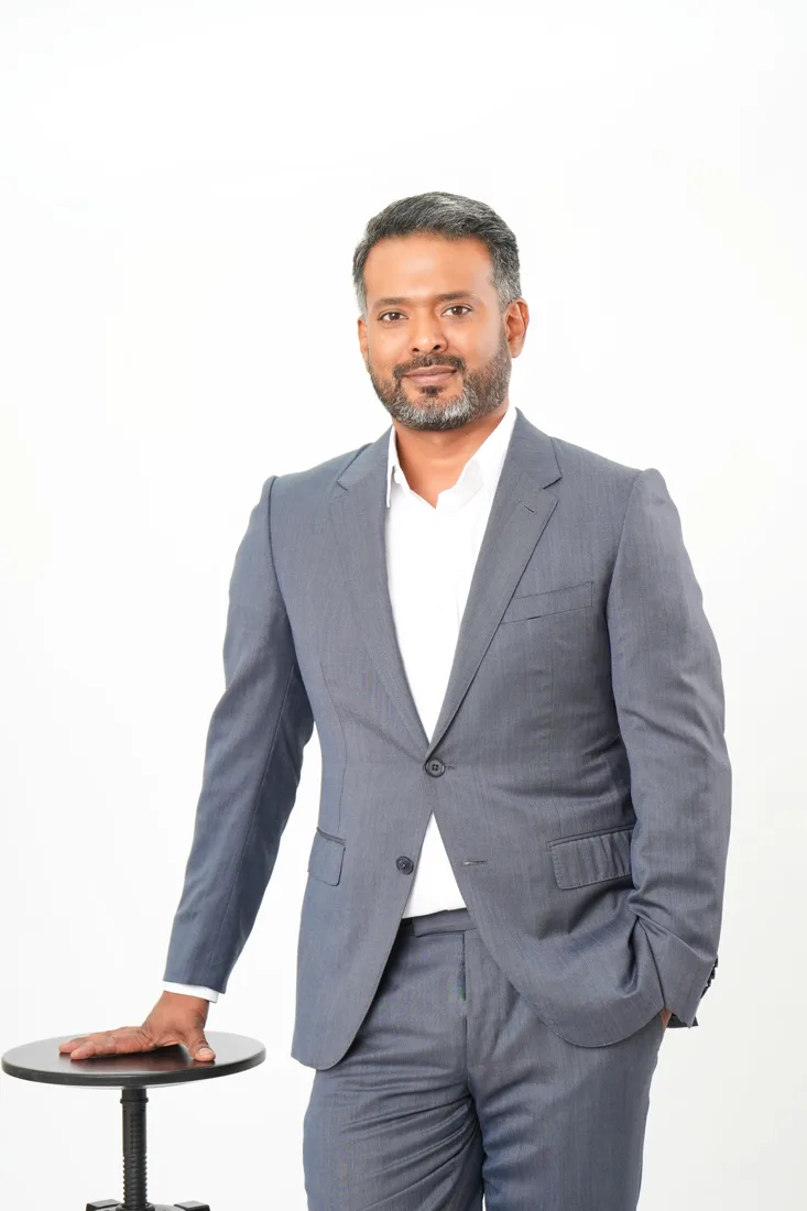 Ash Khan standing beside a small table in a grey suit